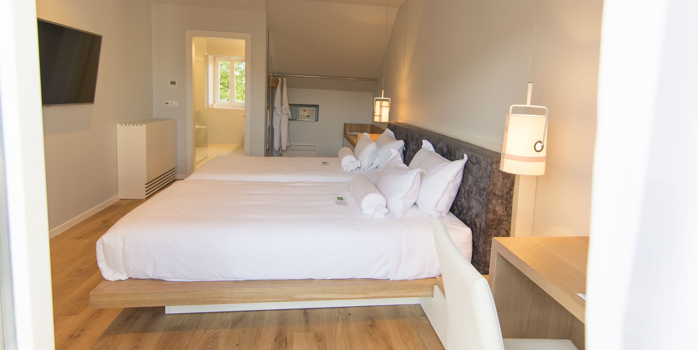A bedroom with two beds, a wooden floor, a TV mounted on the wall, and a bathroom visible through an open door.