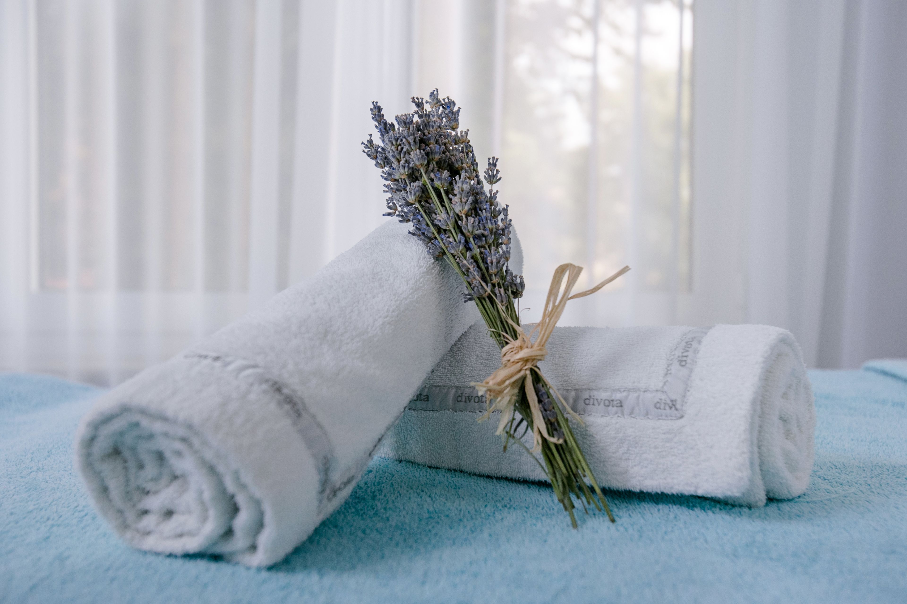 Ein gerolltes weißes Handtuch und ein Bündel Lavendel, zusammengebunden mit einer Schnur, liegen auf einem blauen Handtuch.