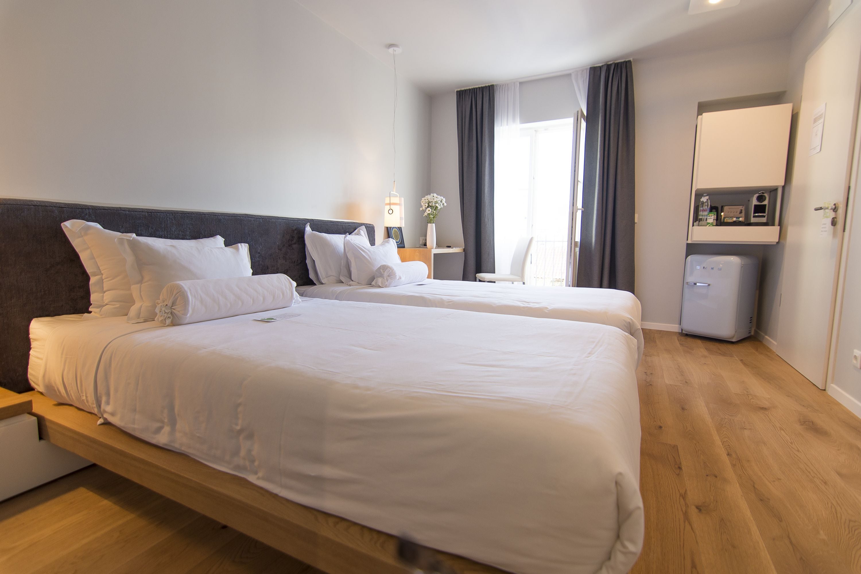 A double bed with white linens, a bedside table with a lamp, a small white chair, and a minibar.
