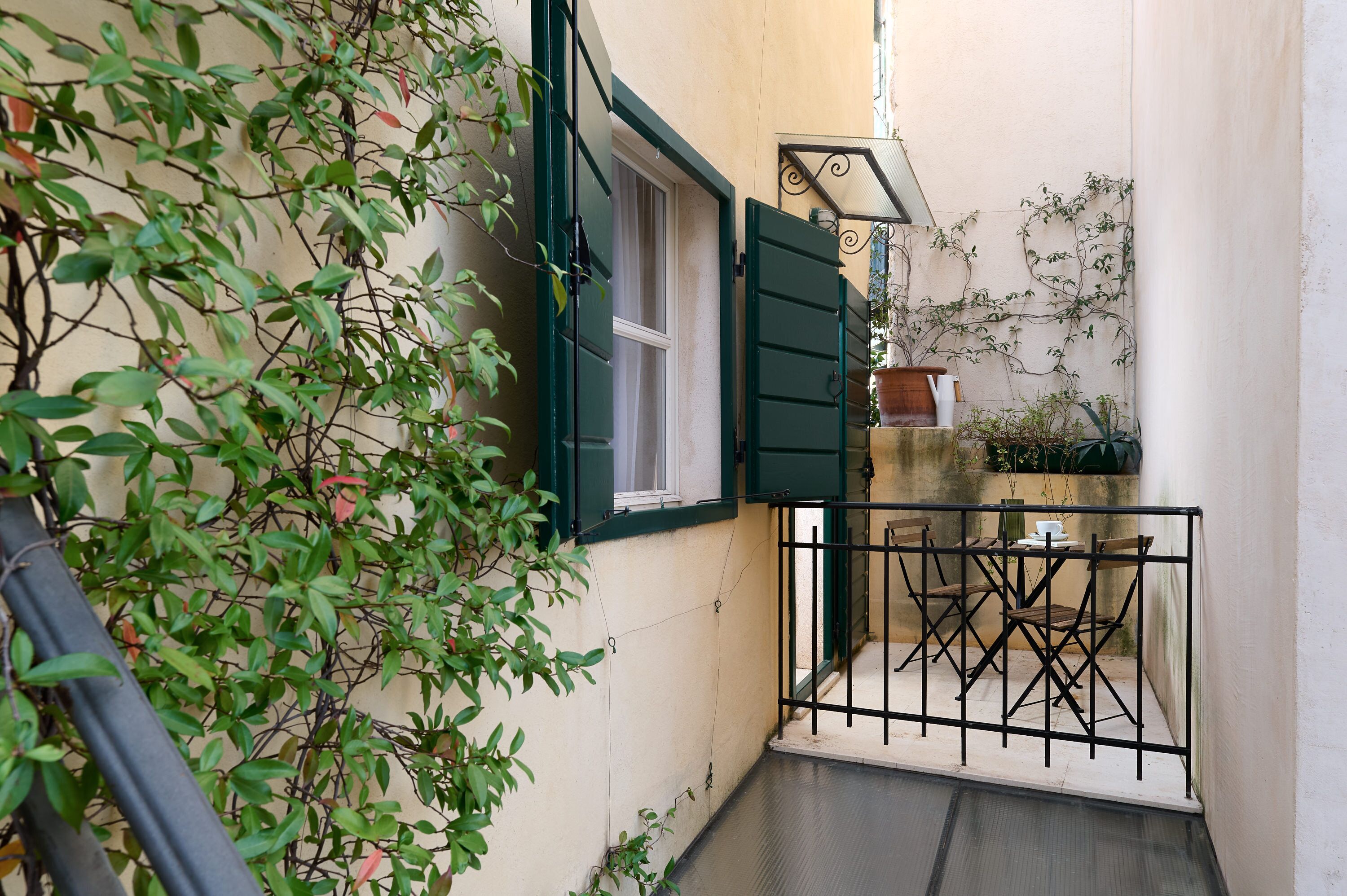 Ein schmales, offenes Raum mit einem kleinen Tisch und Stühlen, einem Fenster mit grünen Fensterläden und Kletterpflanzen an der Wand.