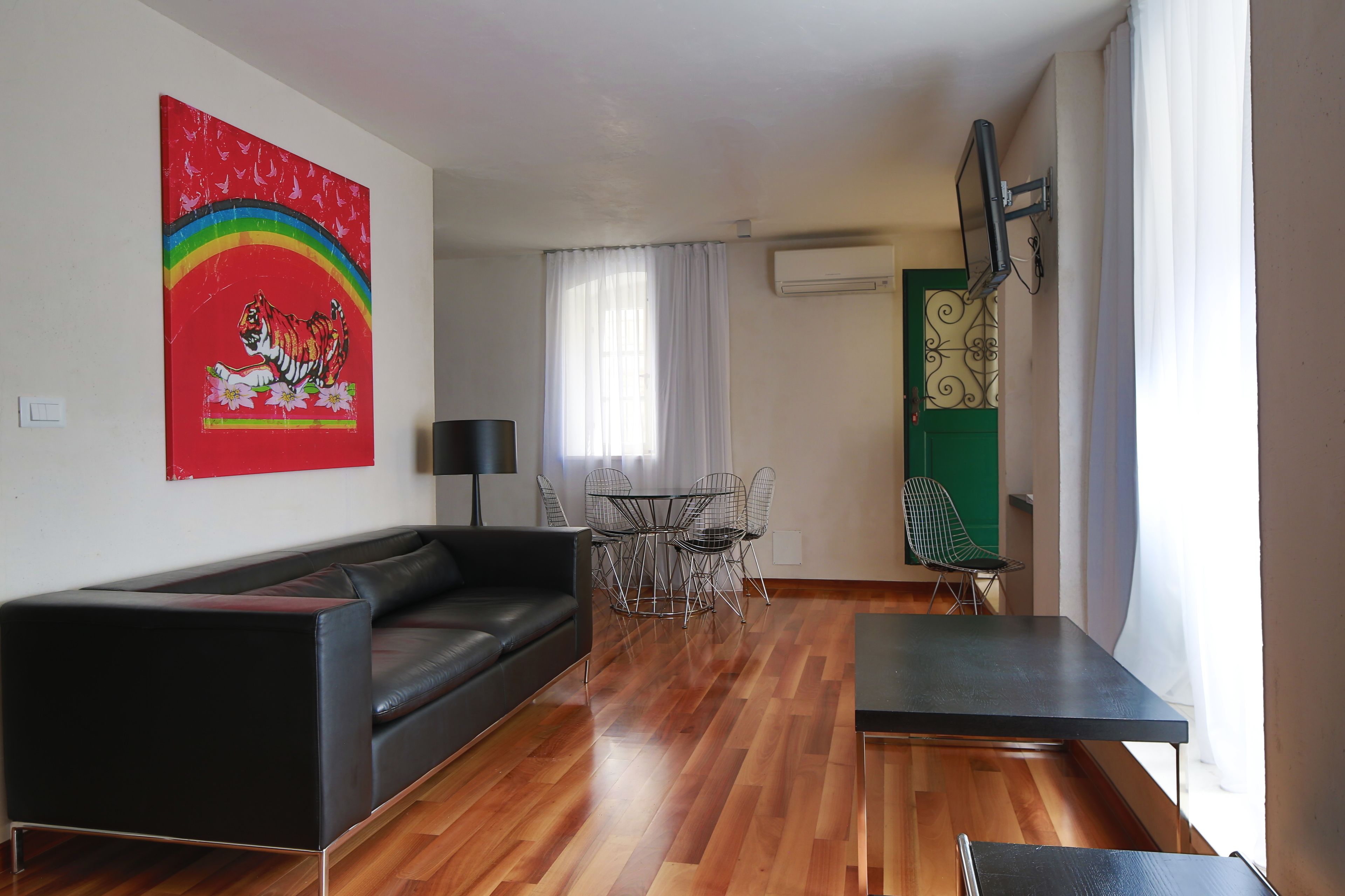 A living room with a black leather sofa, a dining table with chairs, a TV mounted on the wall, and an air conditioning unit.