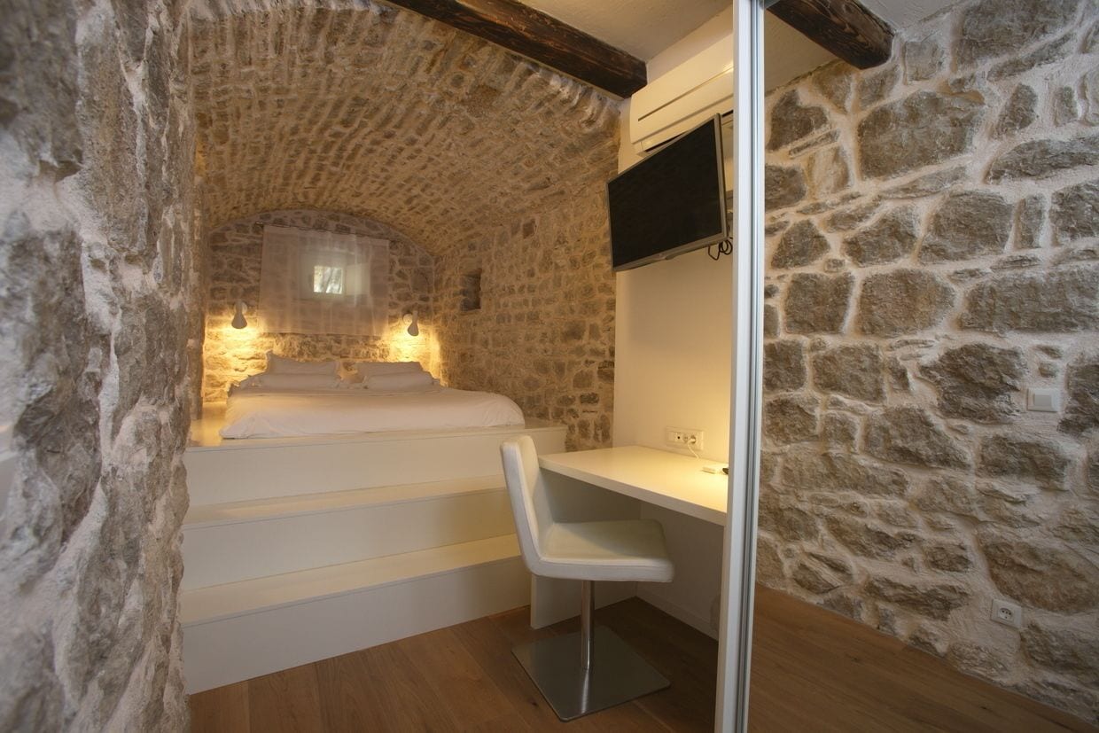 A small, stone-walled room with a bed, a desk, and a chair.