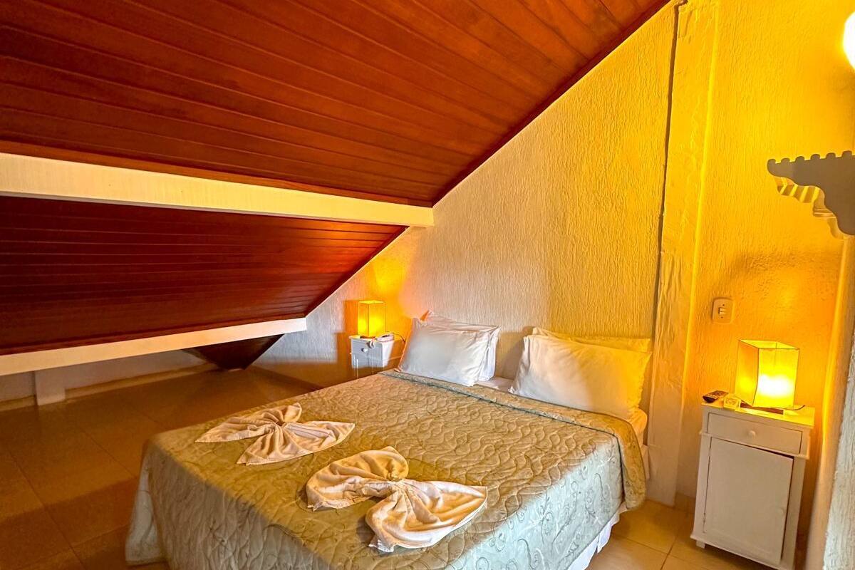 Ein Schlafzimmer mit schrägem Dach, einem Bett mit Steppdecke, zwei Nachttischen mit Lampen und einer an der Wand befestigten Leuchte.