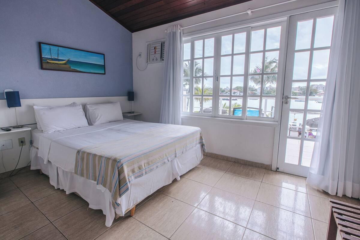 Ein Schlafzimmer mit Bett, Nachttisch, Wandsteckdose, Fenster mit Vorhängen und Blick auf einen Swimmingpool und Palmen.