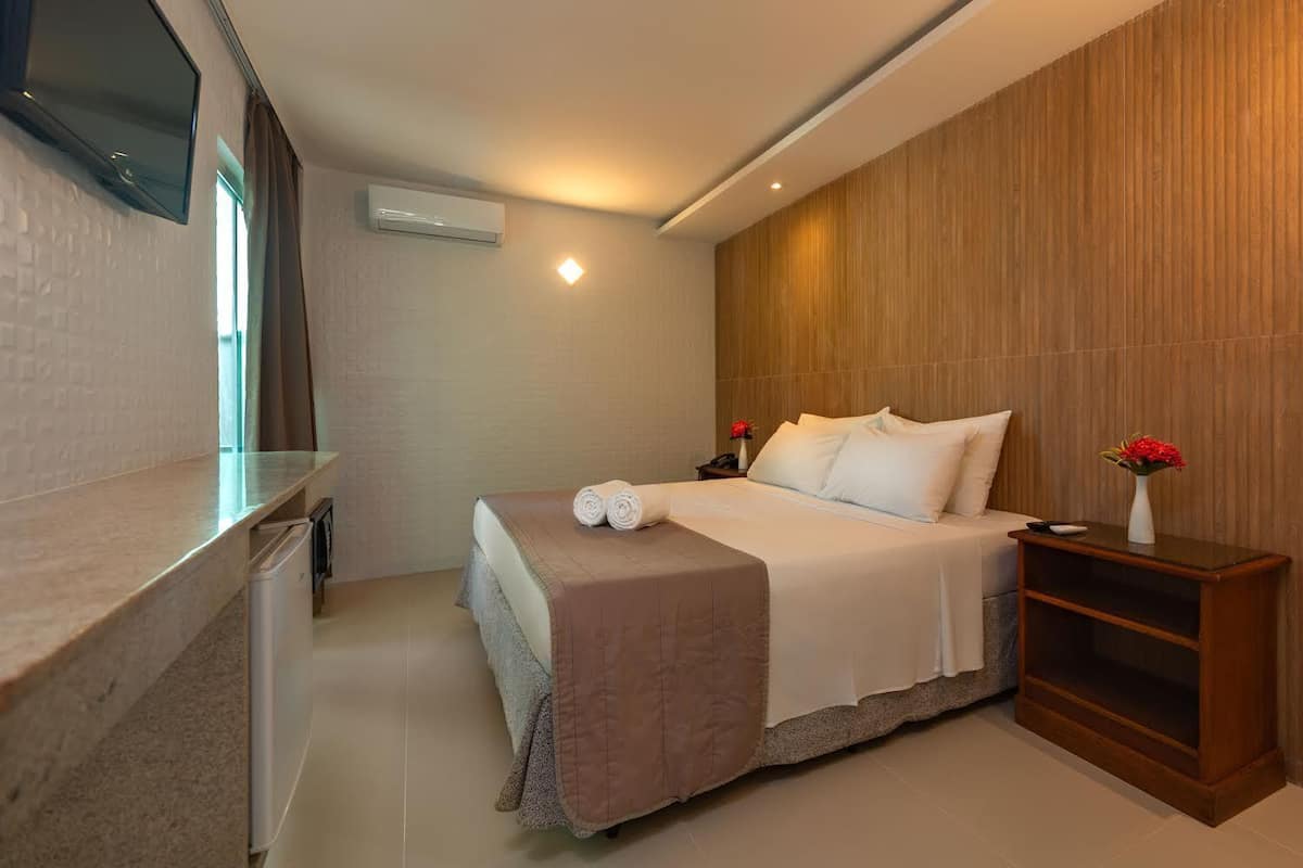A modern hotel room with a bed, a bedside table with a vase of flowers, a flat-screen TV mounted on the wall, and an air conditioning unit.