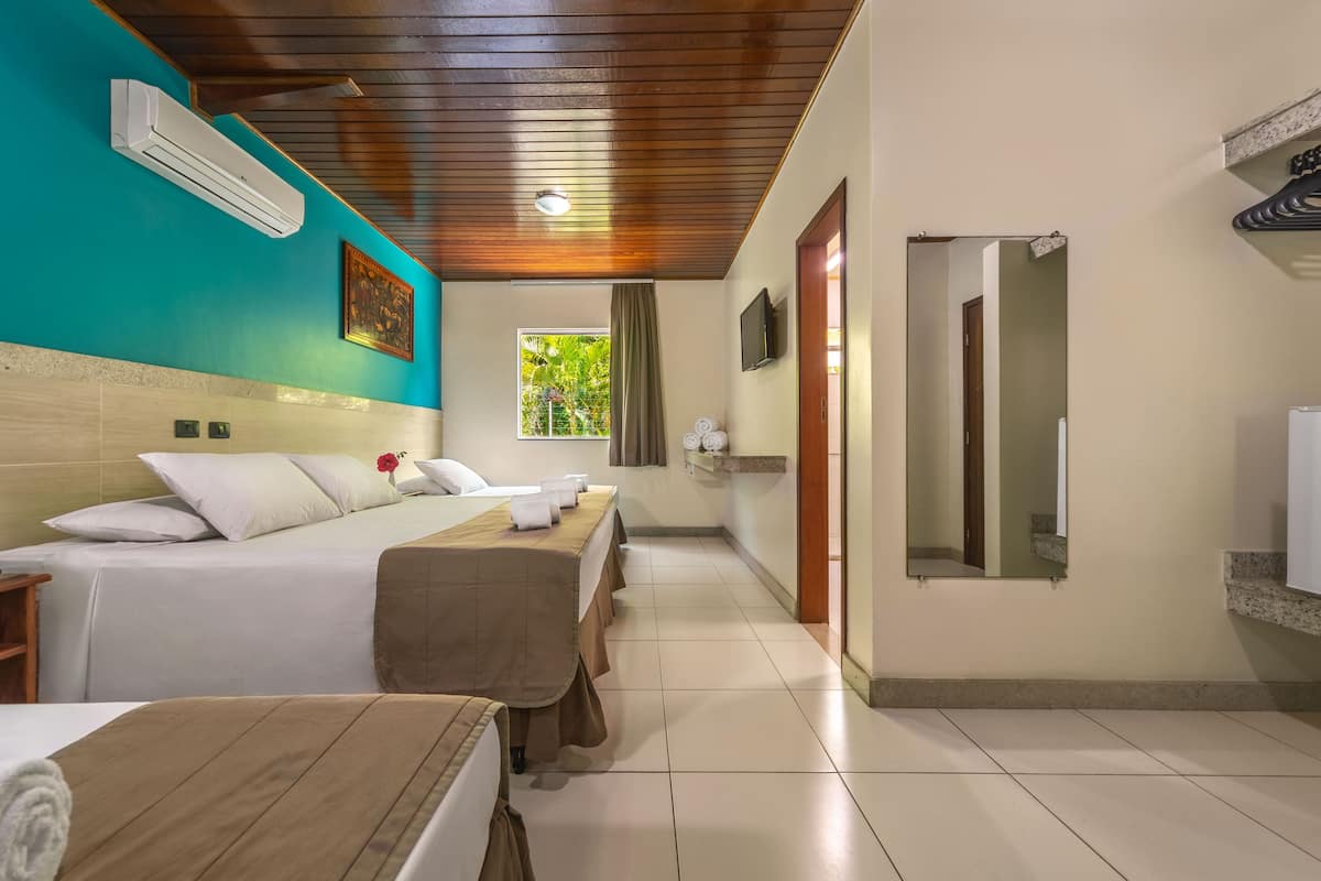 A hotel room with a bed, a wooden headboard, a window with a view of greenery, a mirror, and a bathroom with a sink and towel rack.