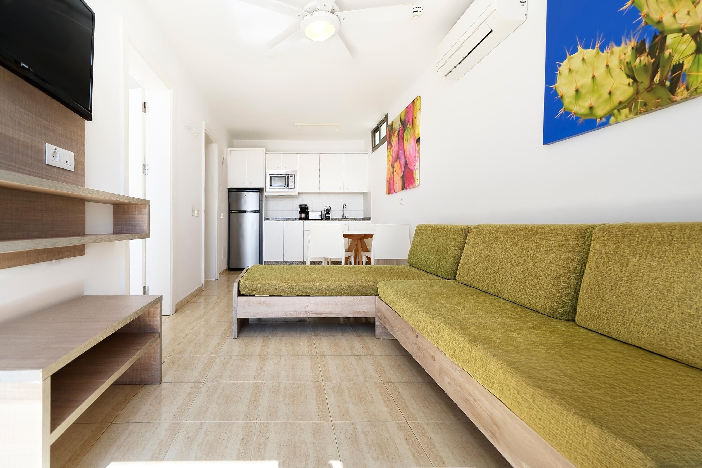 Una sala moderna con un sofá verde, un televisor de pantalla plana, estanterías de madera, una cocina al fondo y un ventilador de techo.