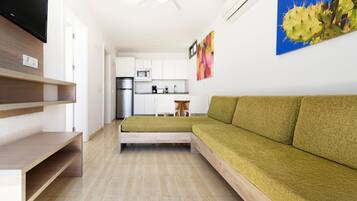 Una sala moderna con un sofá verde, un televisor de pantalla plana, estanterías de madera, una cocina al fondo y un ventilador de techo.