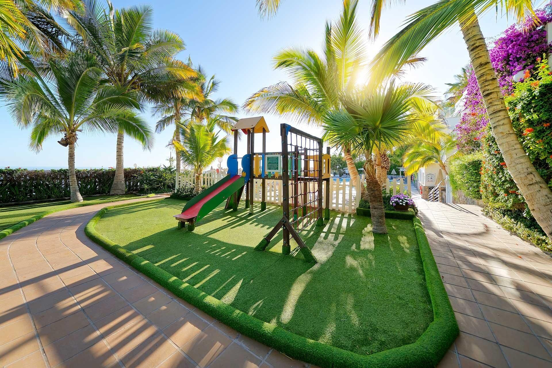 A playground with a slide and climbing structures, surrounded by palm trees and well-maintained grass.