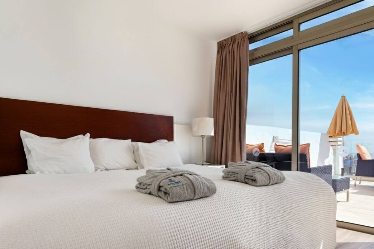 Habitación de hotel con una cama grande, vistas a la ciudad y un balcón con mobiliario exterior.