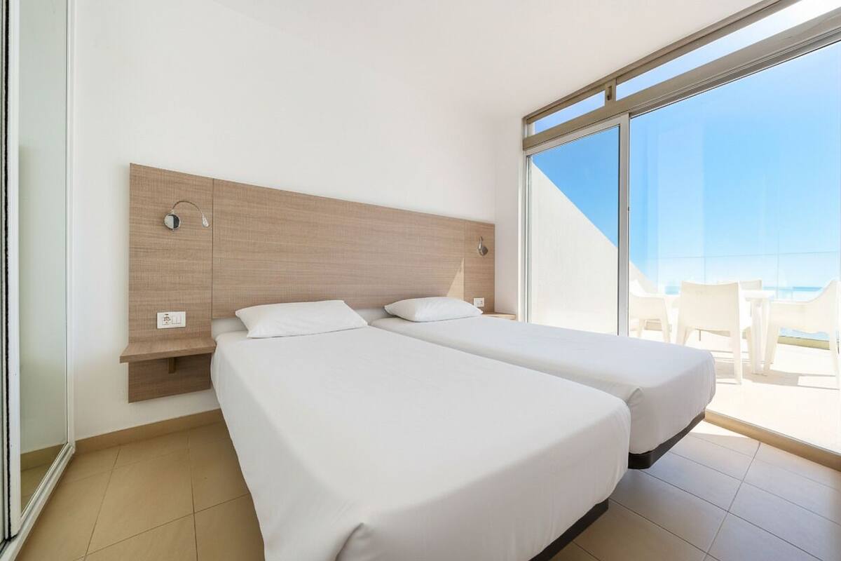 Habitación de hotel con dos camas, cabecero de madera, ventana grande y vista a la playa.