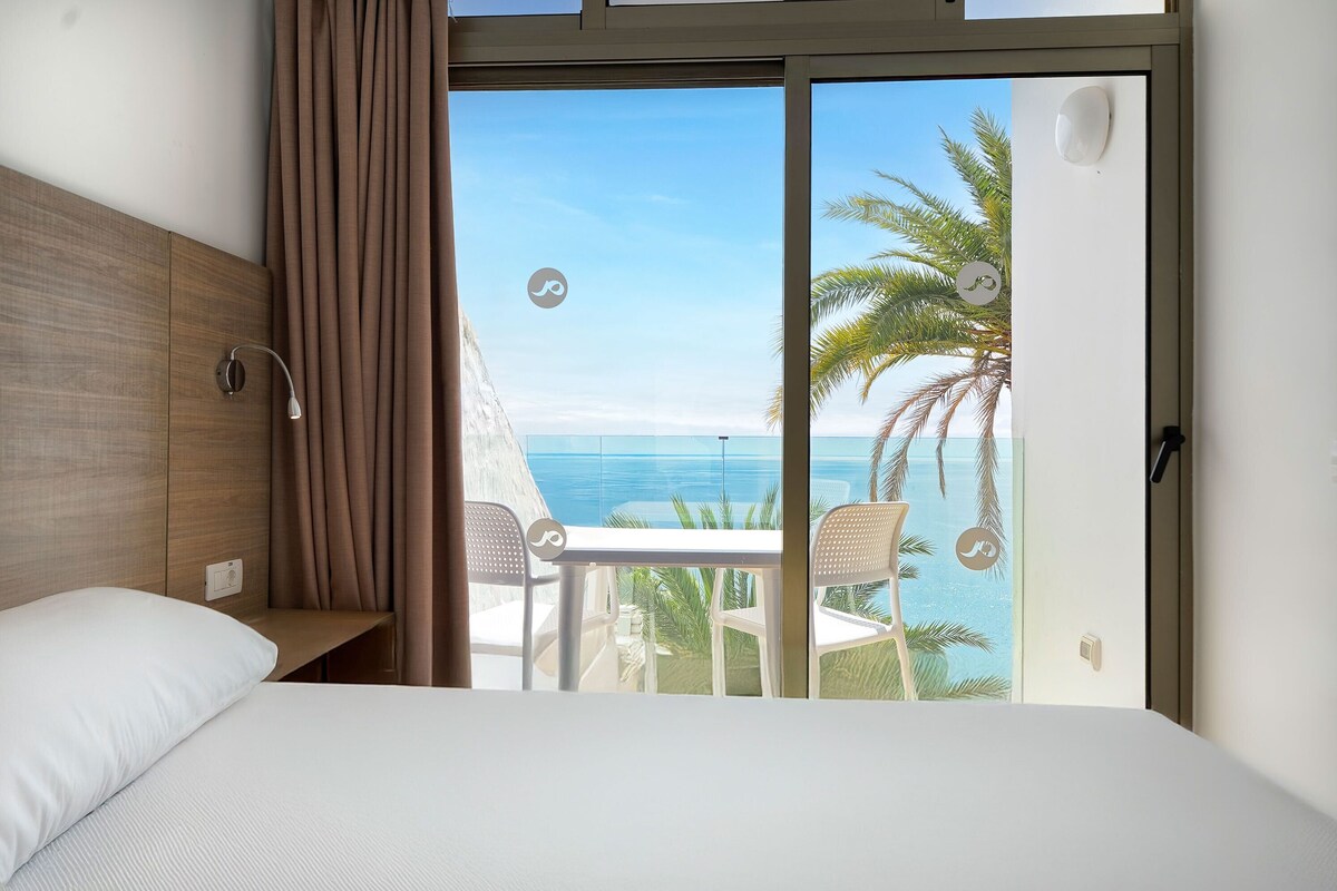 A hotel room with a bed, a small table, and a view of the ocean through a sliding door.