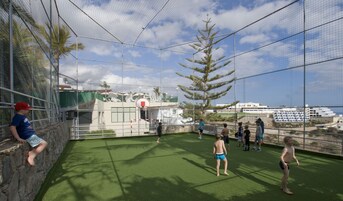 Un área deportiva al aire libre con aro de baloncesto, césped artificial y una vista de un crucero al fondo.