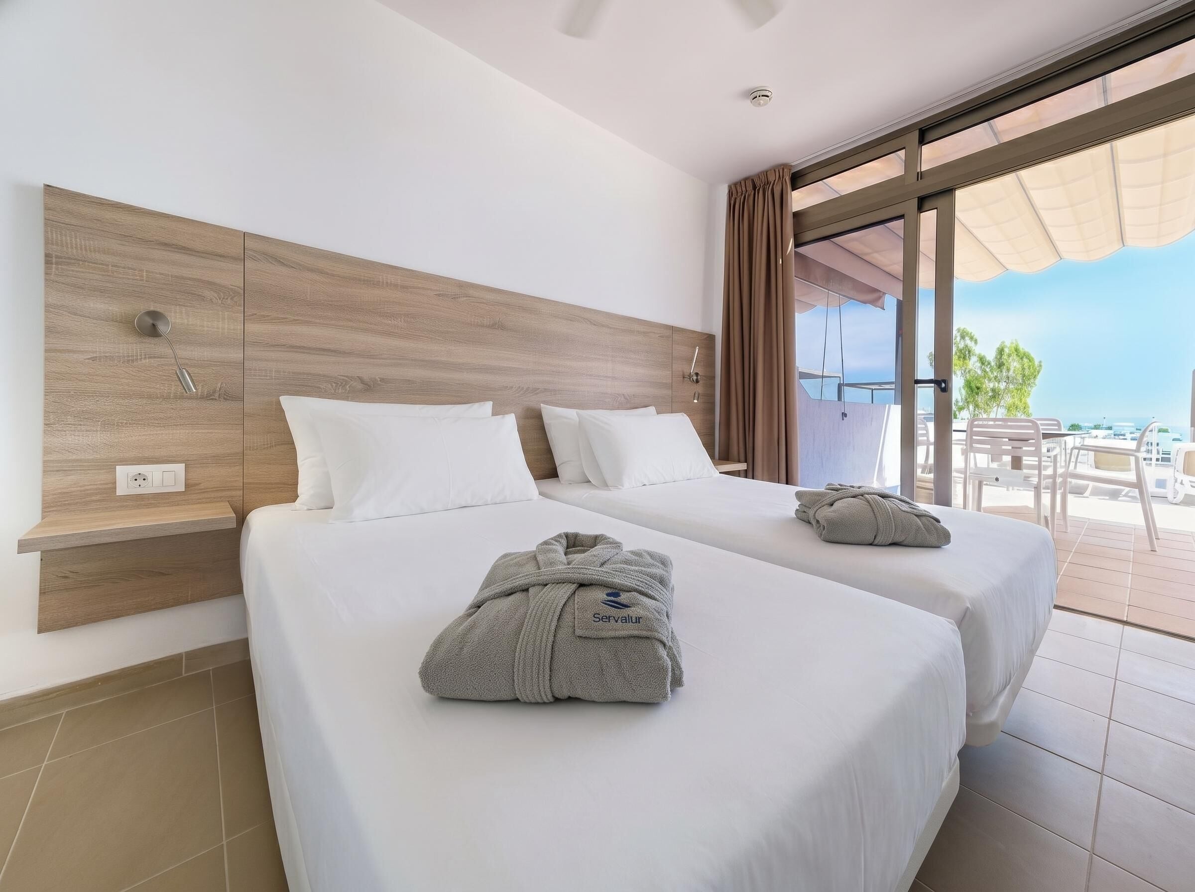 A hotel room with two beds, a wooden headboard, a large window with a view, and a balcony with outdoor seating.