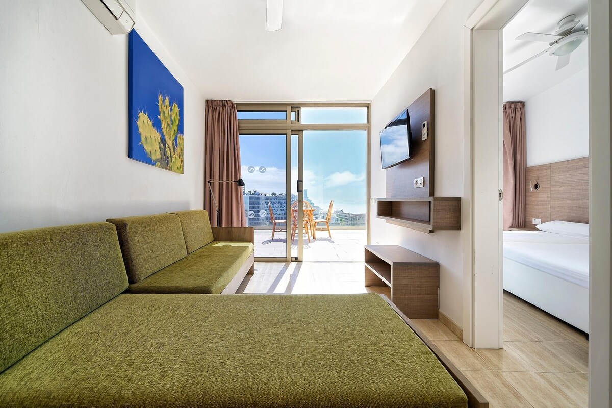 A modern hotel room with a green sofa, a flat-screen TV, a bed, and a city view through the sliding glass door.
