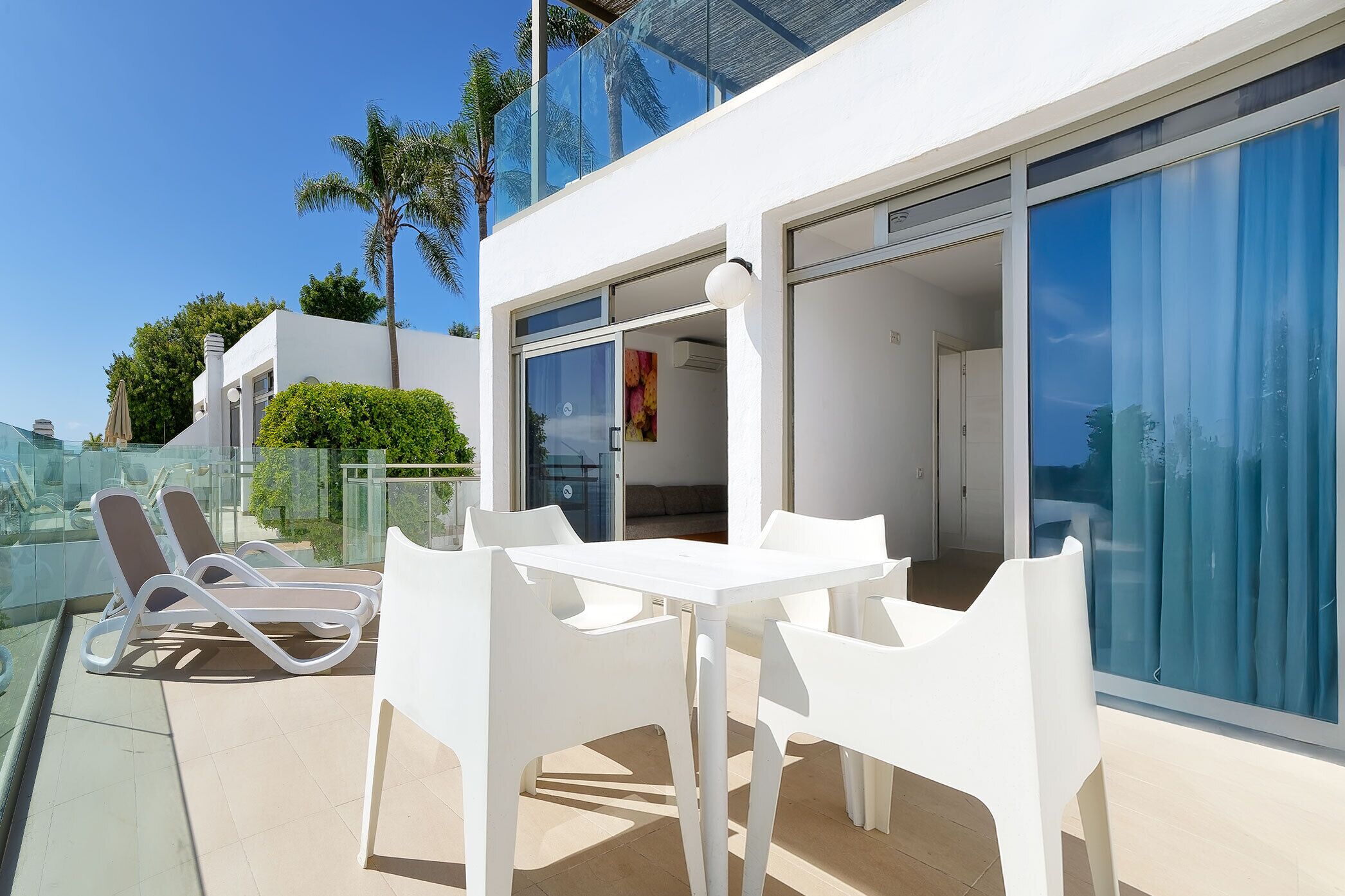 A modern outdoor patio with white furniture, a glass balustrade, and a view of the surrounding area.