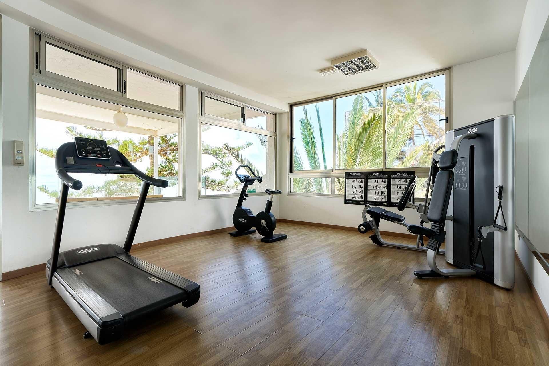 A spacious gym with a treadmill, elliptical, and weight machines, large windows with a view of palm trees.