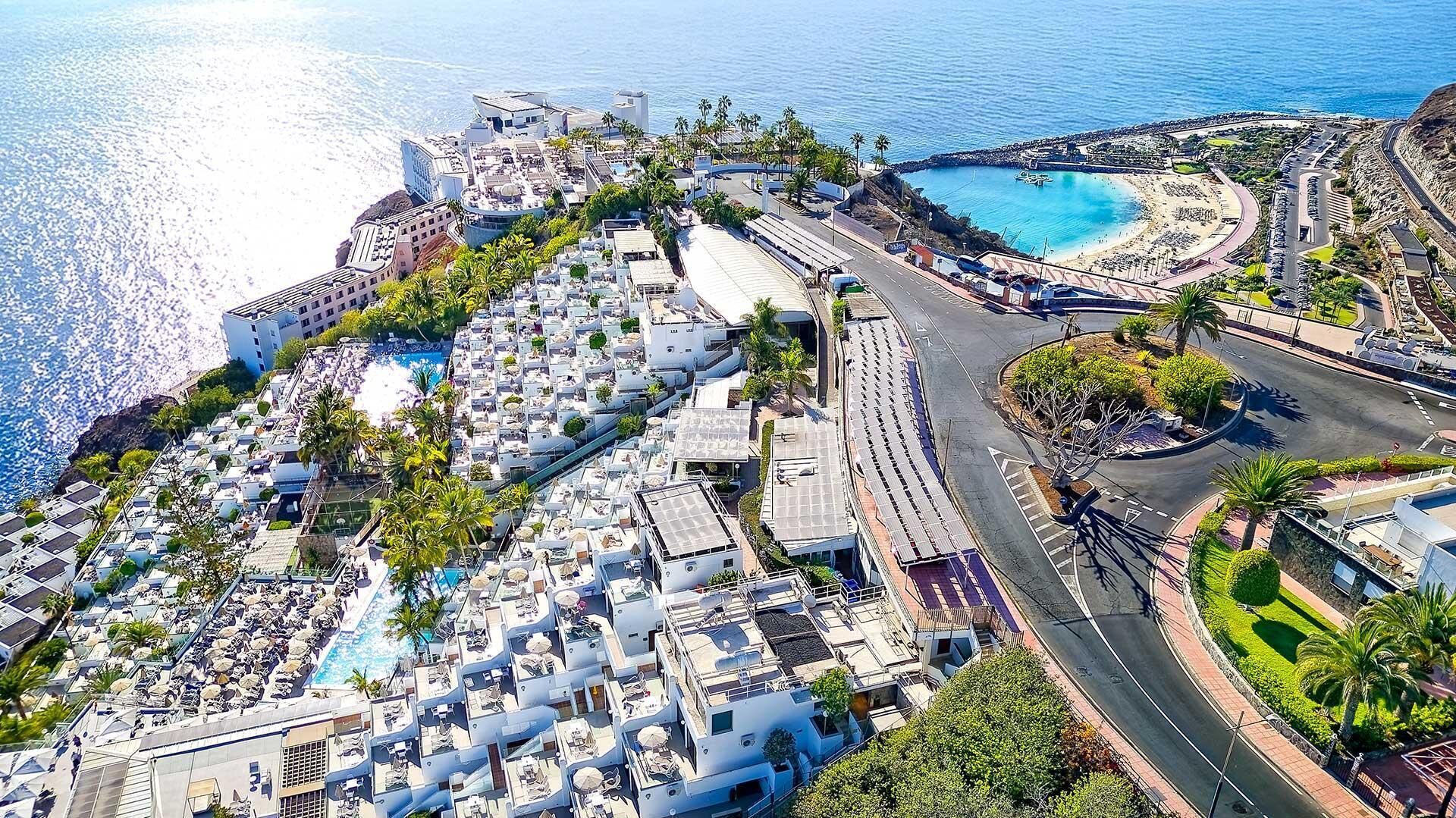Vista aérea de um resort costeiro com vários edifícios, uma grande piscina e uma estrada sinuosa que leva a uma praia.