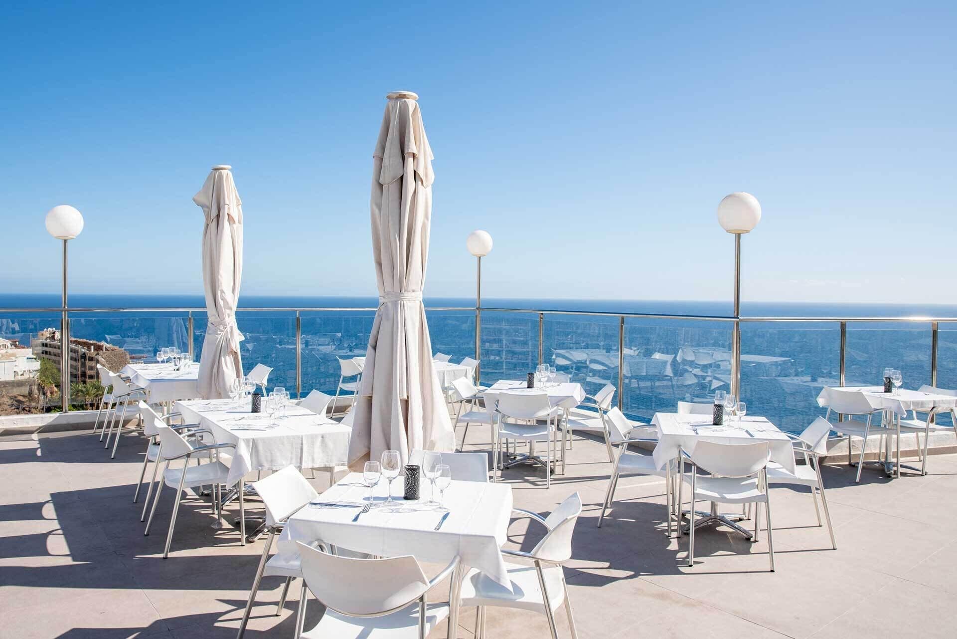 An outdoor dining area with tables and chairs, white umbrellas, and a view of the ocean.
