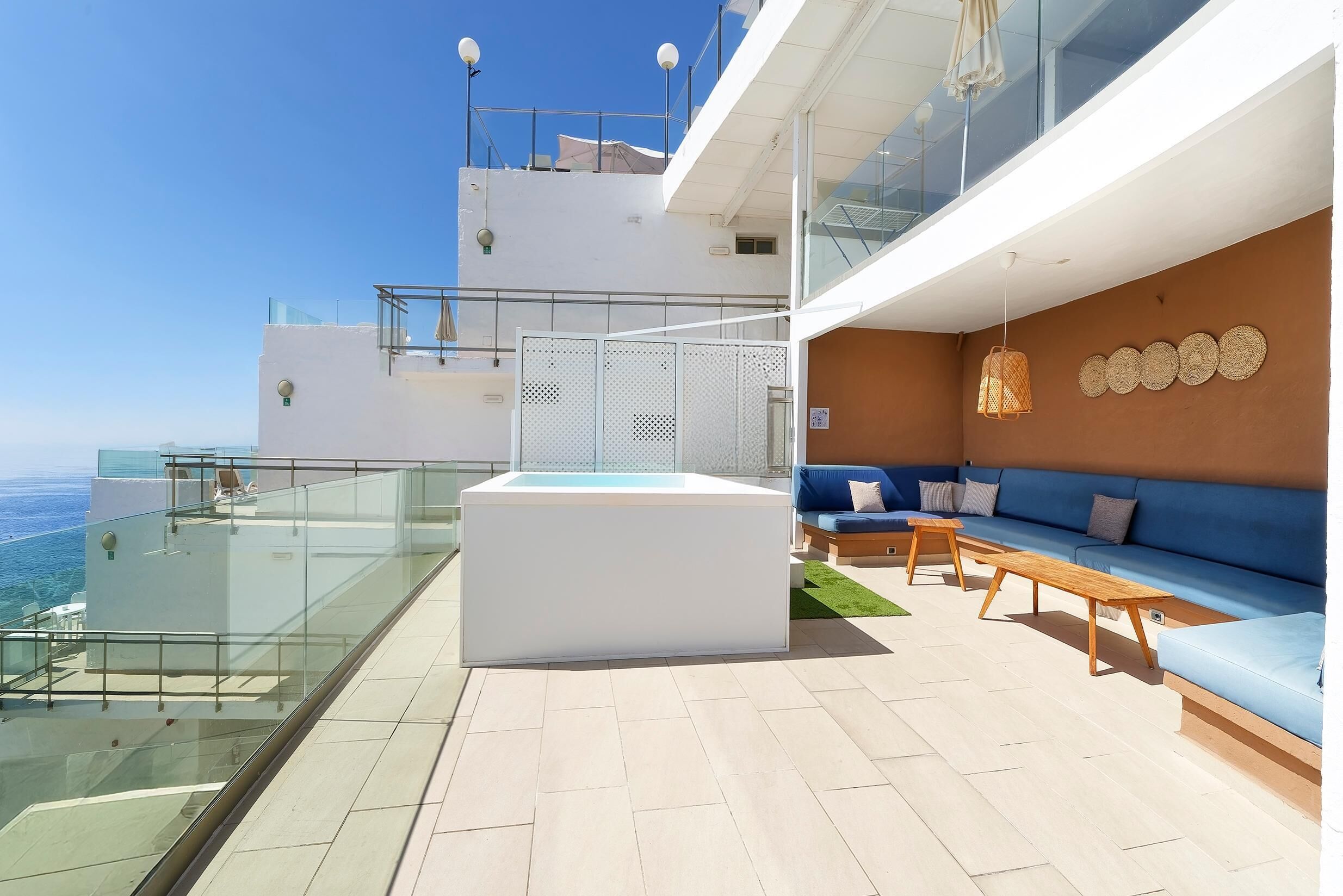 A modern outdoor terrace with a glass balustrade, a blue sofa, wooden tables, and a white structure.