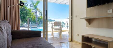 A modern living room with a sofa, a TV mounted on the wall, and a sliding glass door leading to an outdoor area with a pool and chairs.