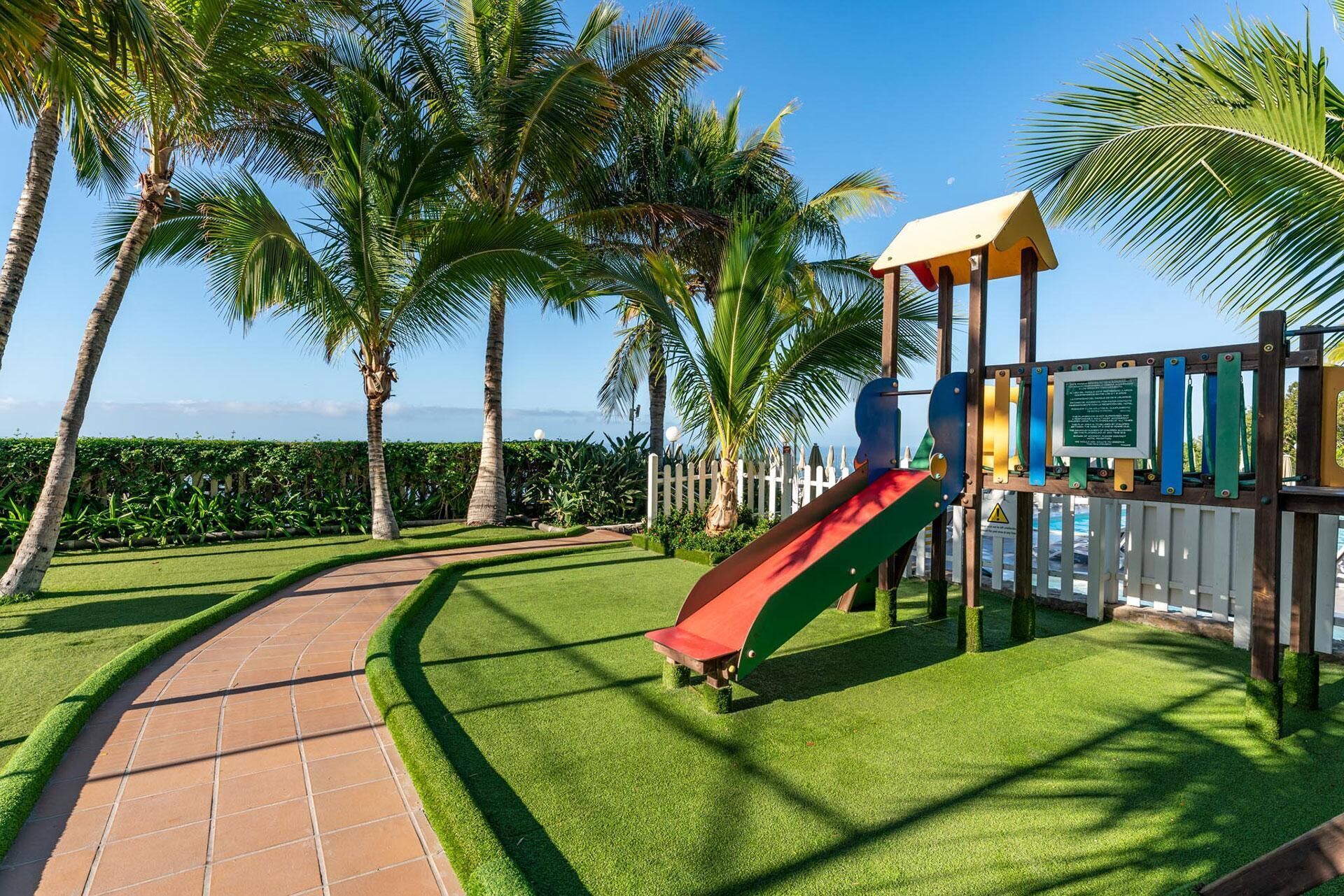 A playground with a slide, surrounded by palm trees and a white picket fence.