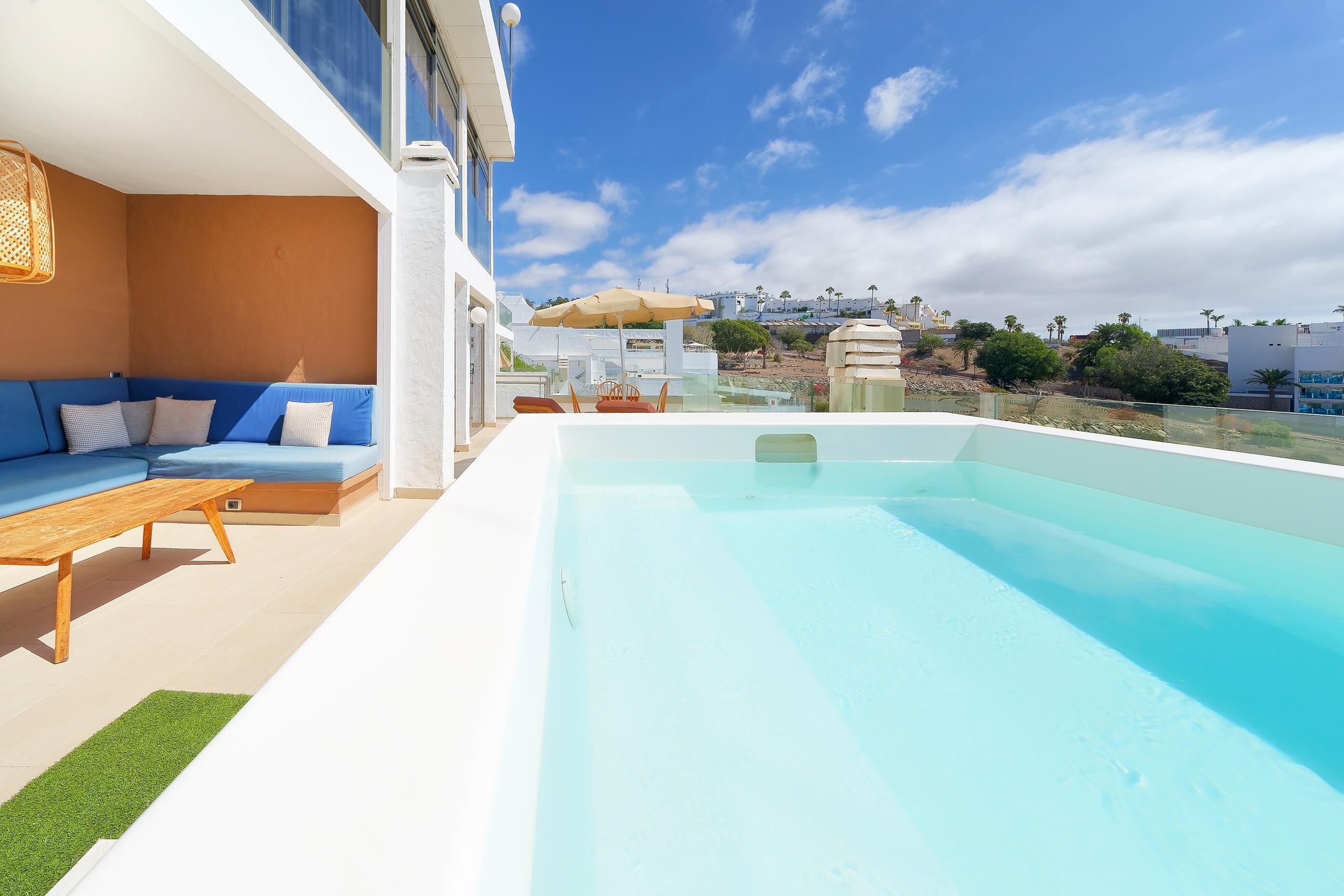 Un área de piscina en la azotea con un sofá azul y una mesa de madera, una vista despejada del paisaje urbano y un edificio blanco al fondo.