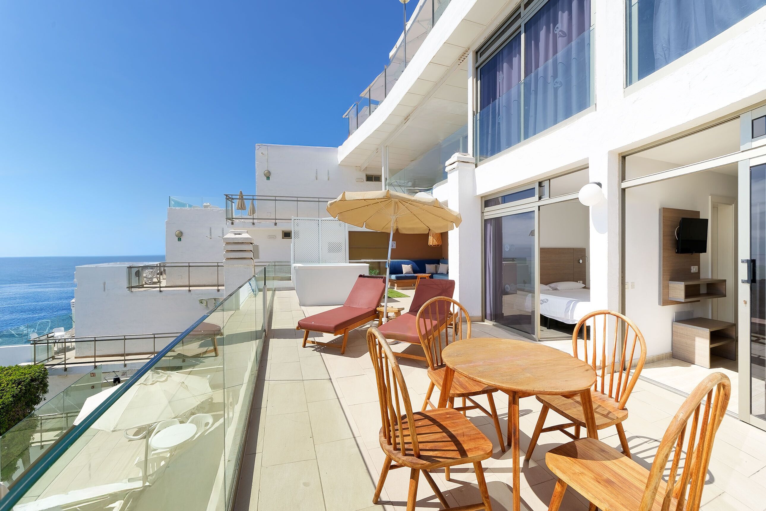A balcony with a glass railing overlooking the ocean, furnished with wooden chairs and a table, and a beige umbrella.