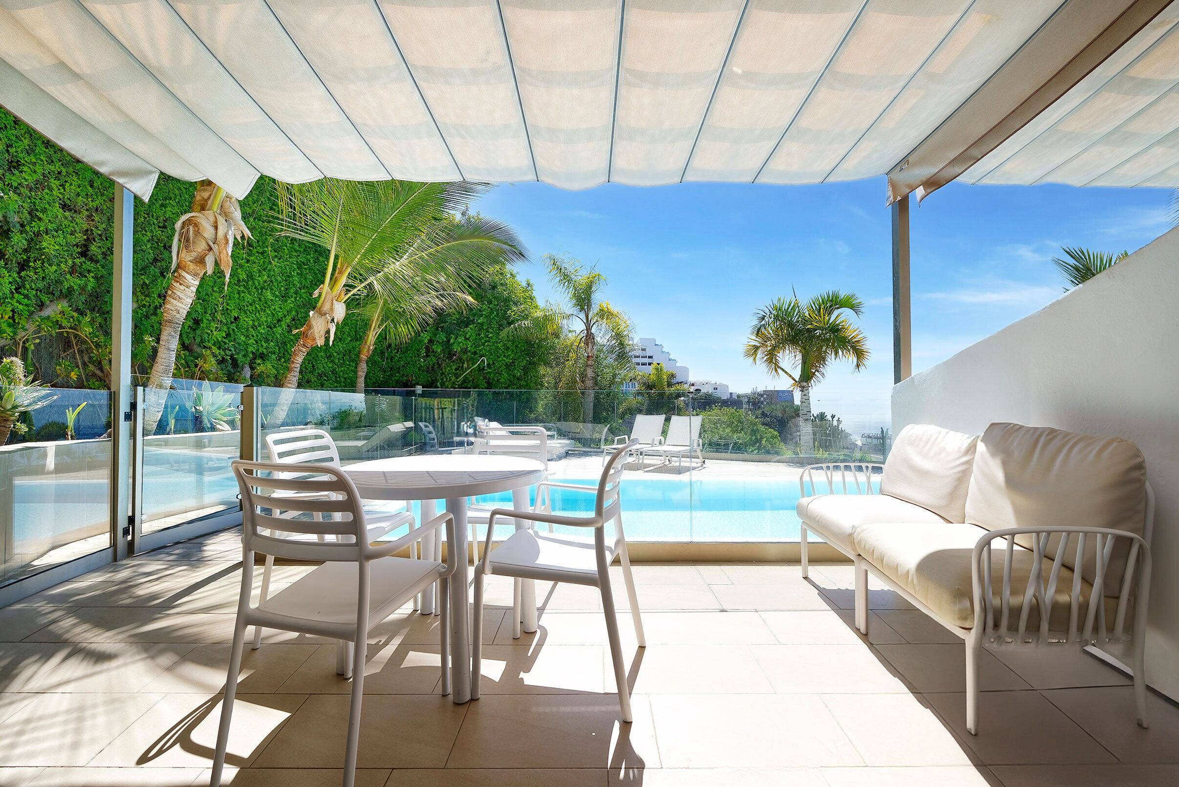 A sunlit outdoor area with a pool, white furniture, and a view of greenery and buildings.