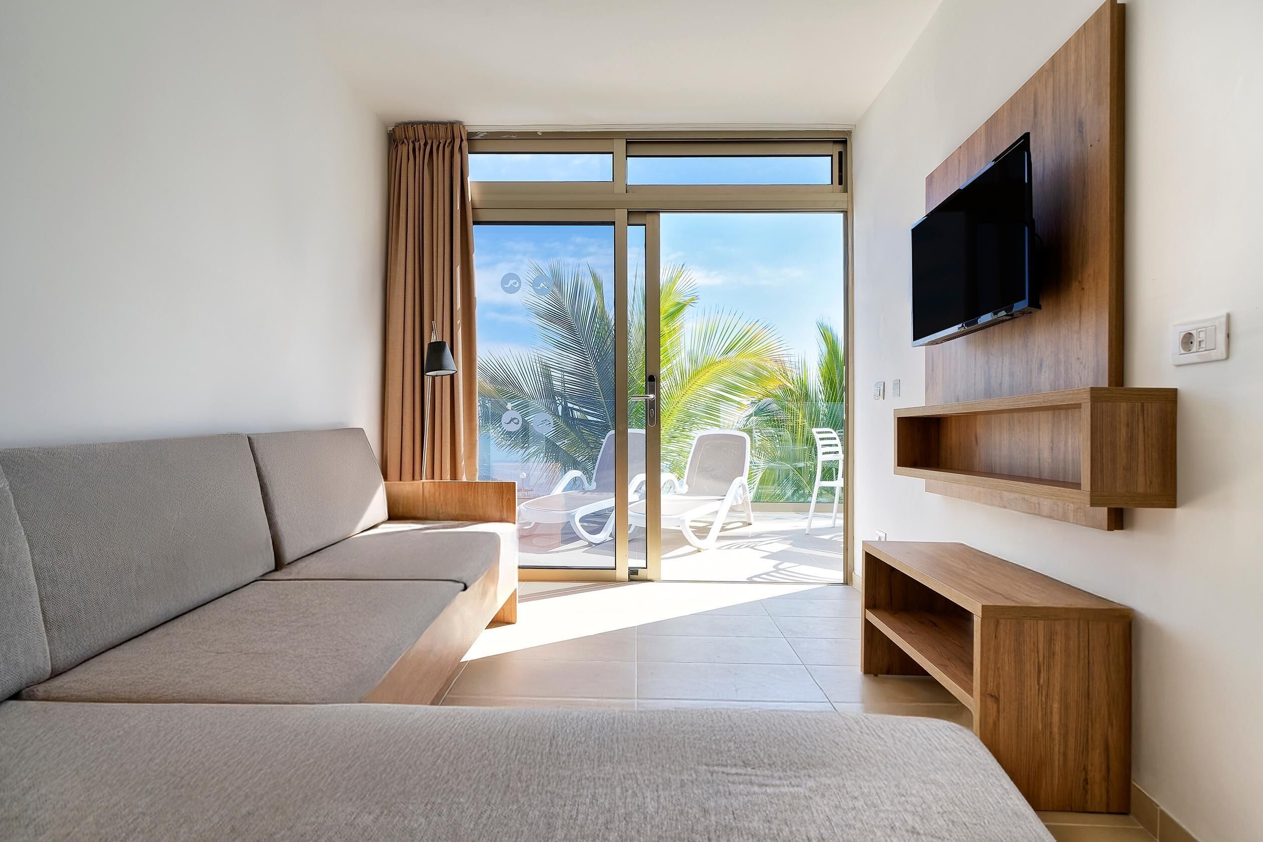 A modern living room with a sofa, a TV mounted on the wall, and a sliding glass door leading to an outdoor area with chairs.