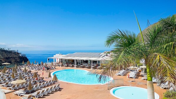 A resort pool area with sun loungers and umbrellas, a covered seating area, and a view of the ocean. - Servatur Altamadores (Mogan)