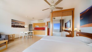 Habitación de hotel con una cama grande, un escritorio, una silla, un sofá, un ventilador de techo y vista a la playa por la ventana.