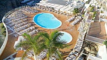 Vista aérea de un complejo turístico con una gran piscina central, múltiples tumbonas y una palmera.