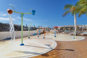 Parque acuático con múltiples atracciones, incluyendo una fuente grande y varias cascadas. Se observan sillones de descanso y sombrillas al fondo, lo que sugiere un ambiente de resort.