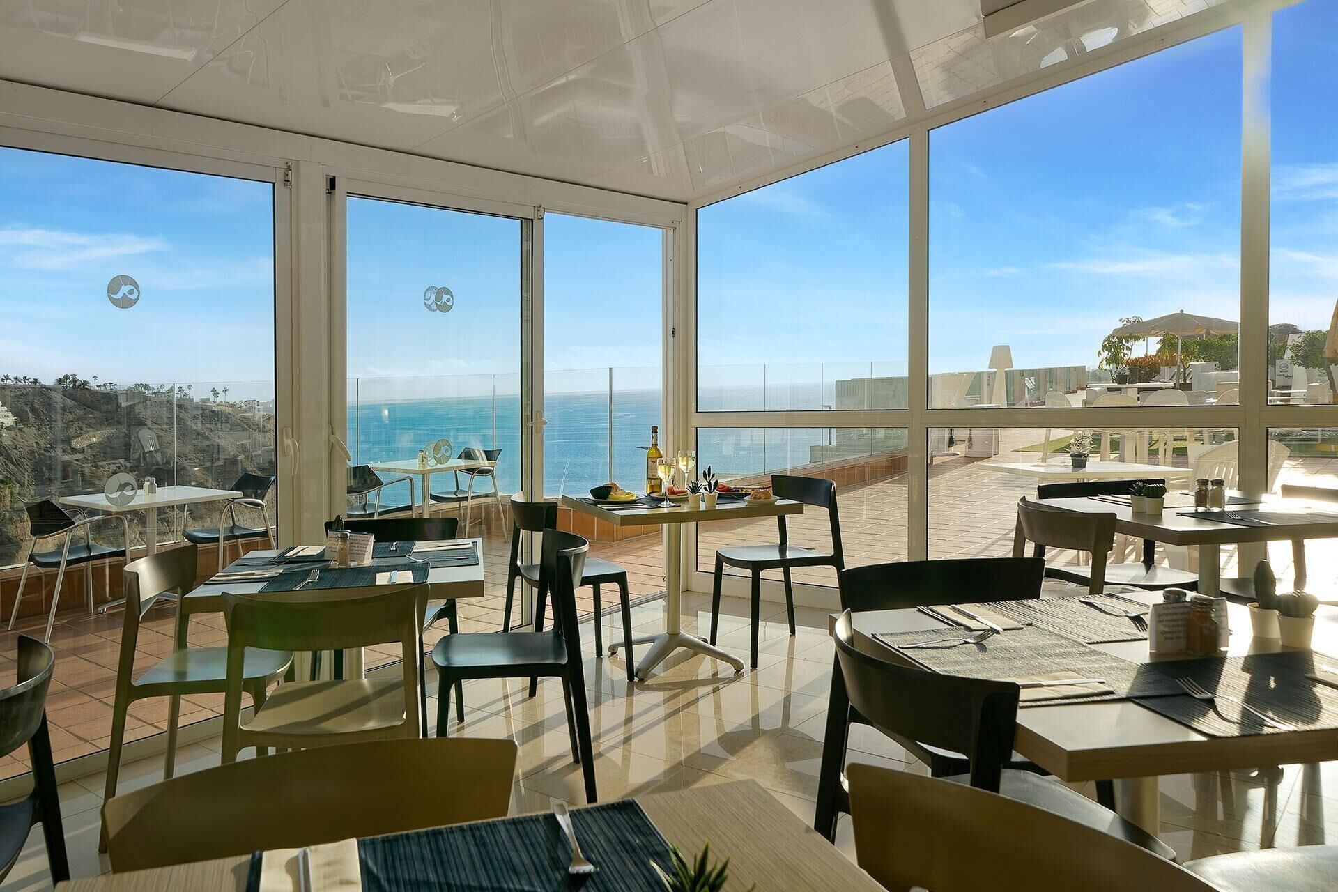 Un restaurante con amplios ventanales que ofrecen vistas al océano, mesas y sillas dispuestas para comer, y una terraza con asientos al aire libre.