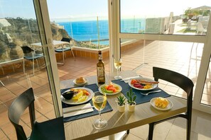 Una mesa de comedor con platos de comida, una botella de vino y copas en un balcón con vistas al océano.