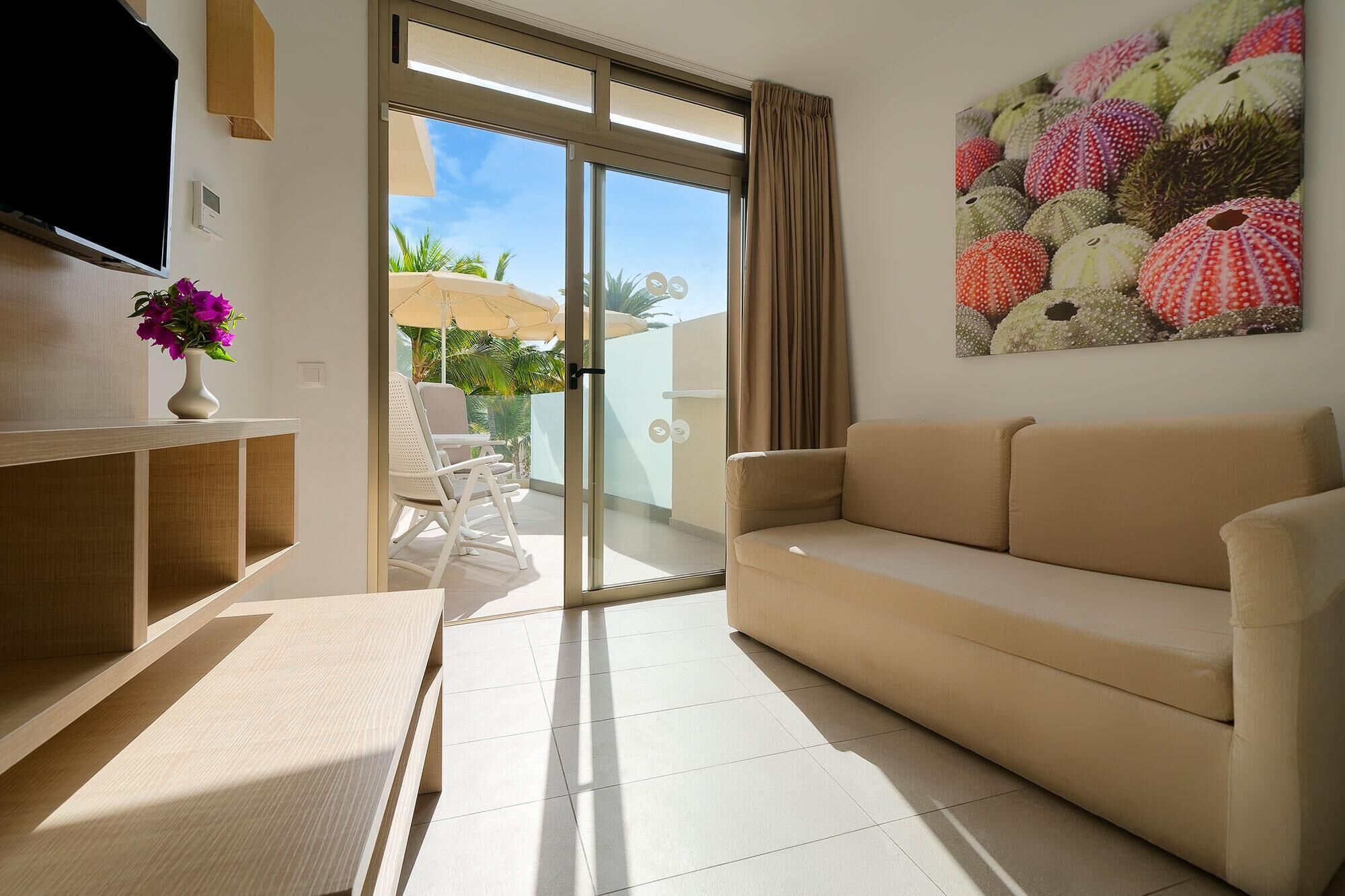Un salon moderne avec un canapé beige, un téléviseur à écran plat, un vase blanc orné de fleurs et une porte-fenêtre coulissante donnant sur un espace extérieur avec une terrasse et une vue sur des palmiers.