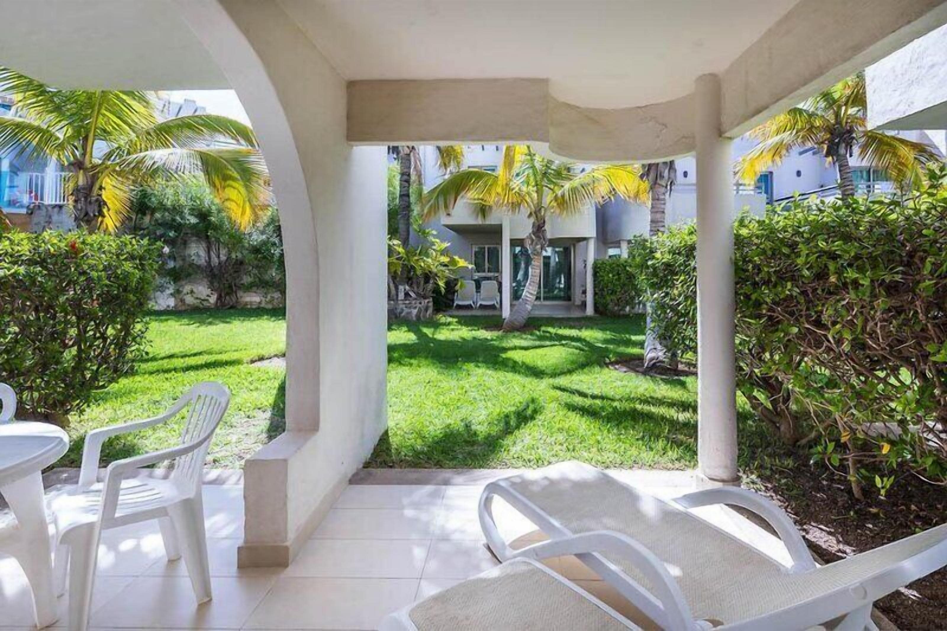 A covered outdoor area with white lounge chairs and a small table, surrounded by greenery and palm trees.