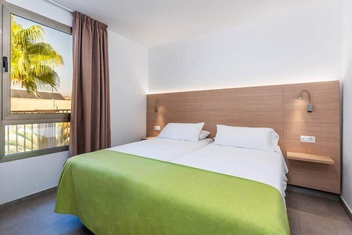 Une chambre d’hôtel avec un lit, une fenêtre avec des rideaux, une vue sur des palmiers et une tête de lit en bois.