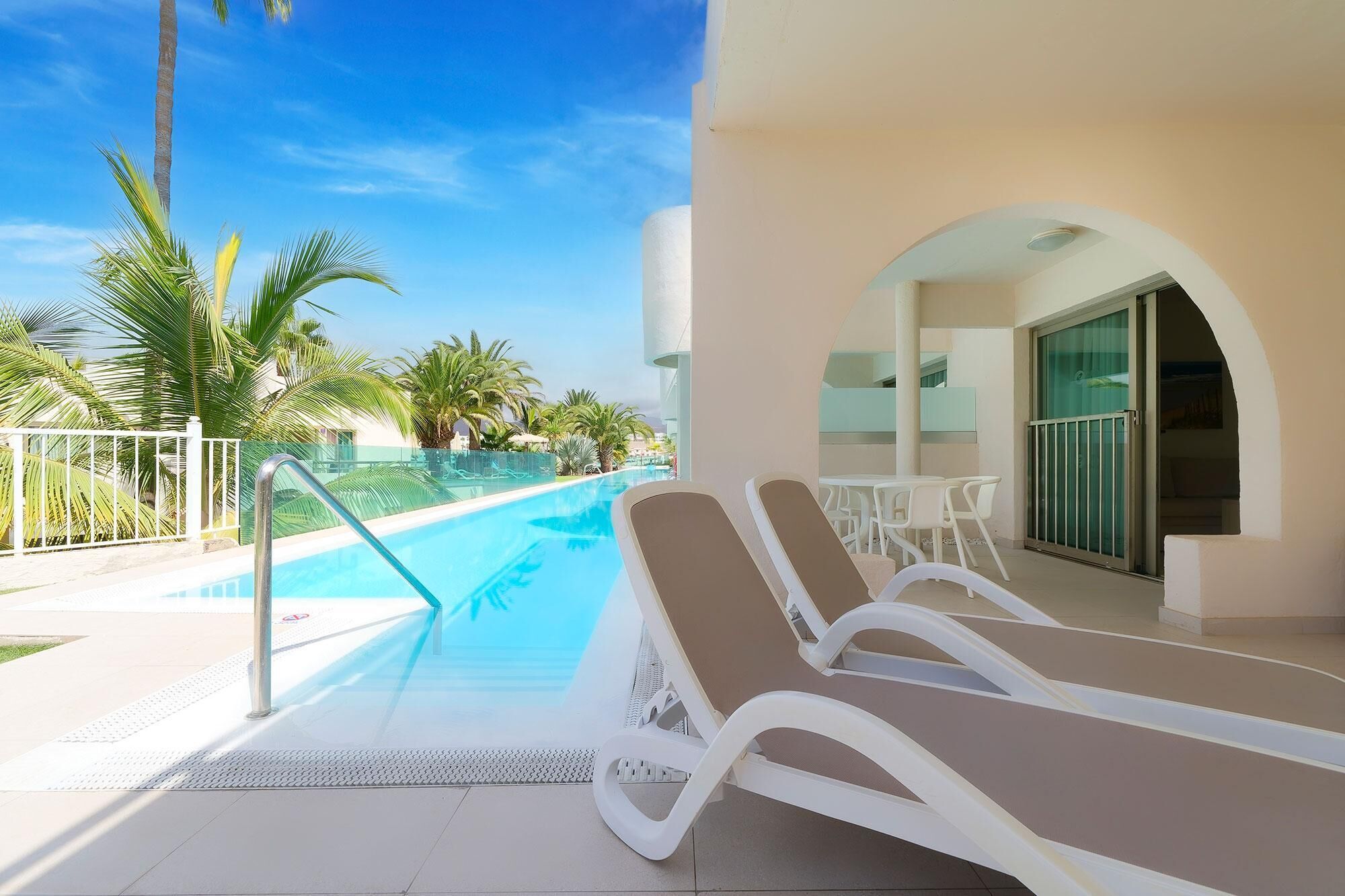 A poolside area with lounge chairs, a glass-enclosed patio, and palm trees.