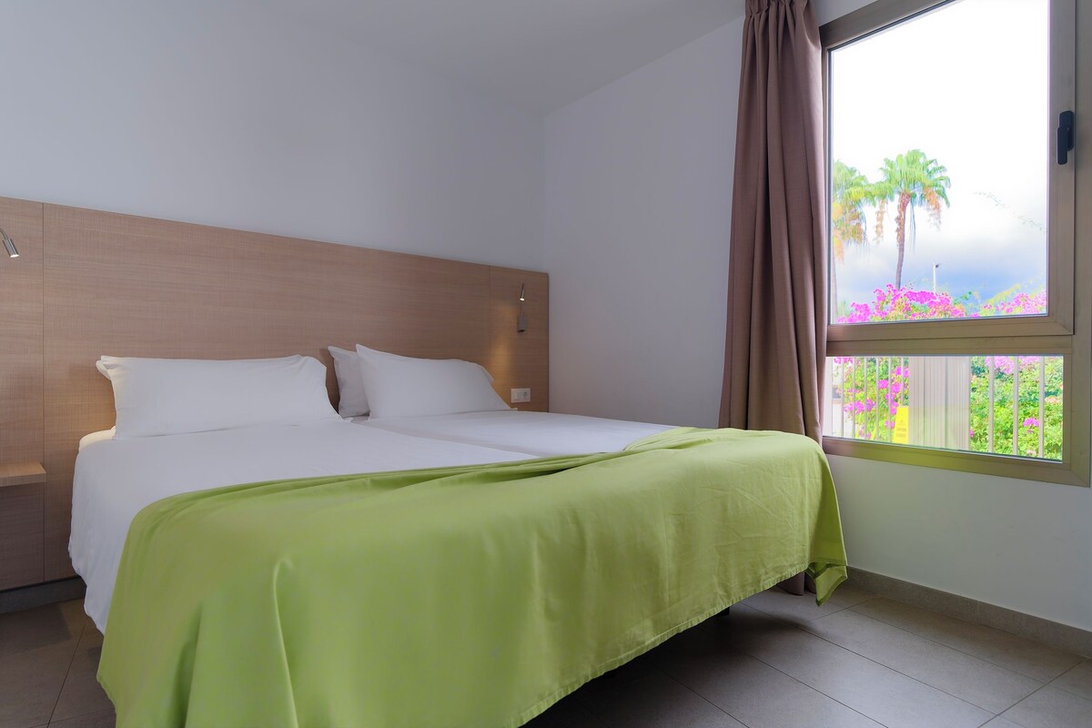 A hotel room with a bed, a window with a view of palm trees and flowers, and a curtain.