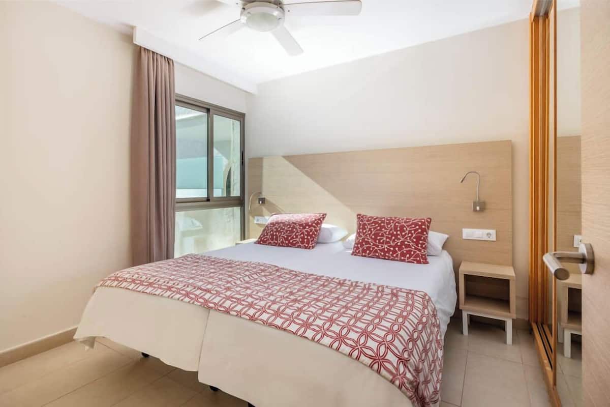 Une chambre d’hôtel avec un lit, une table de chevet, une fenêtre avec des rideaux, un ventilateur de plafond et une tête de lit en bois.