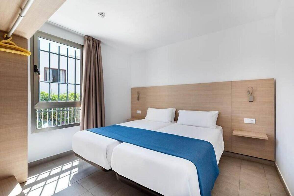 Une chambre d’hôtel avec un lit double, une fenêtre avec des rideaux, une tête de lit en bois et une petite étagère.