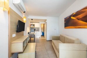 Uma sala de estar moderna com uma TV de tela plana, um sofá, uma mesa de centro e uma área de cozinha ao fundo.