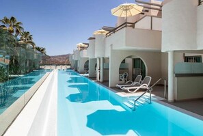 Una piscina con sillas de descanso y sombrillas, rodeada de palmeras y un cielo despejado.