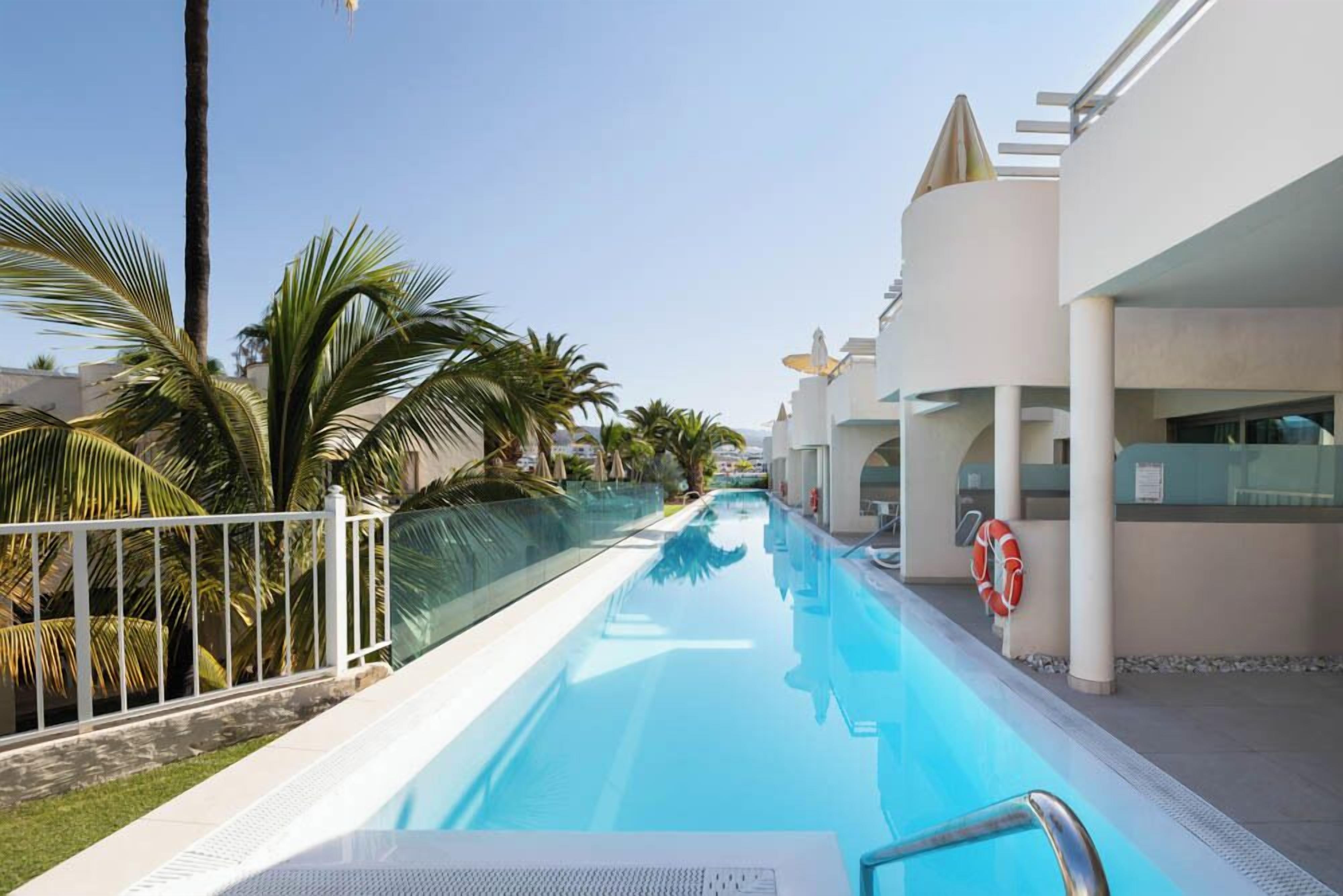 A swimming pool with a clear blue water, surrounded by palm trees and modern buildings.