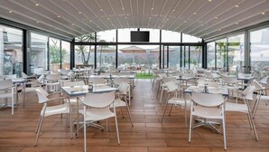 A spacious indoor dining area with modern white chairs and tables, large windows offering views of the outdoors, and a ceiling with linear lighting fixtures. - Servatur Edén & Buganvillas (Mogan)
