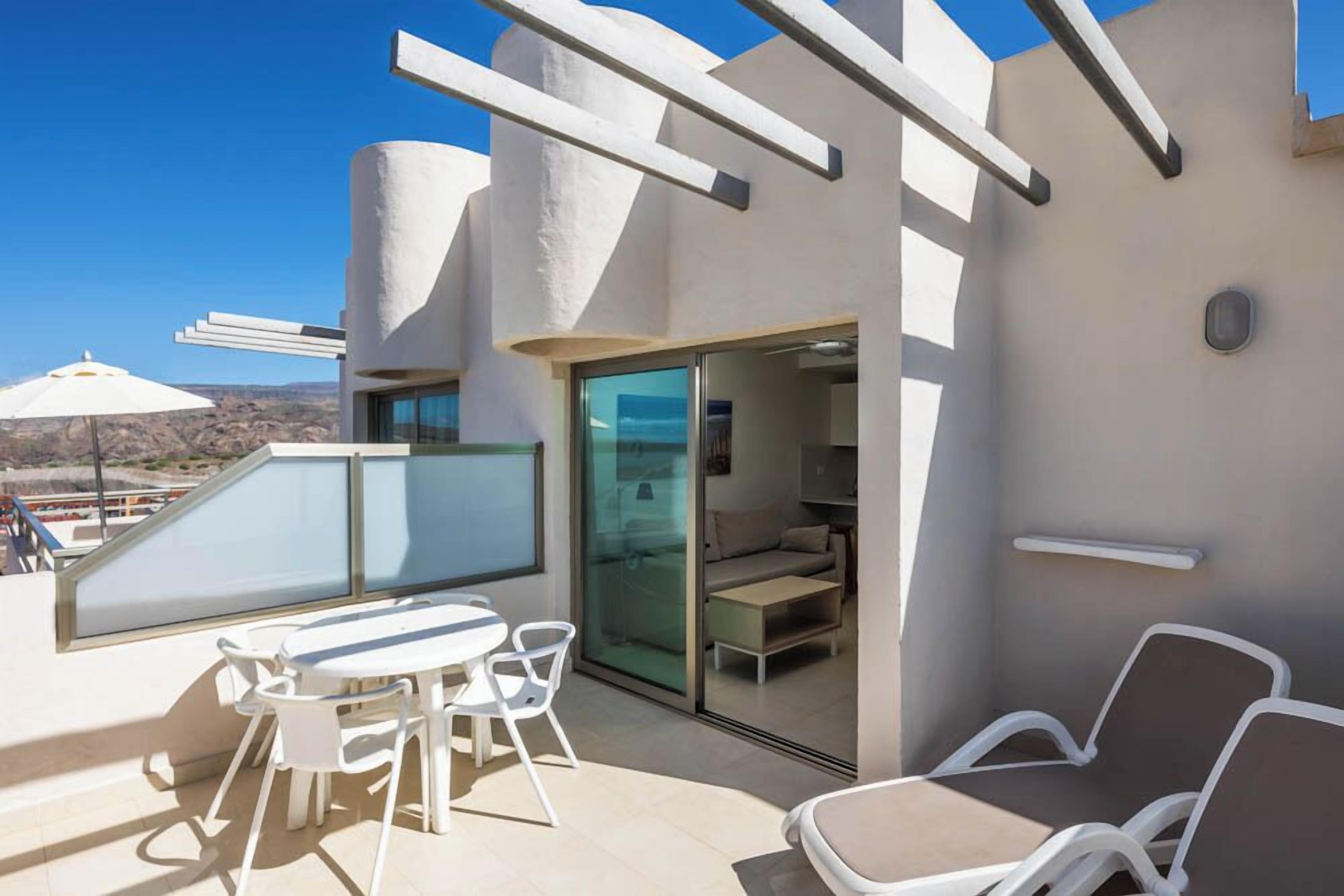 Une terrasse extérieure moderne, dotée d’une porte vitrée donnant sur un espace intérieur, de meubles blancs et offrant une vue dégagée sur le paysage.
