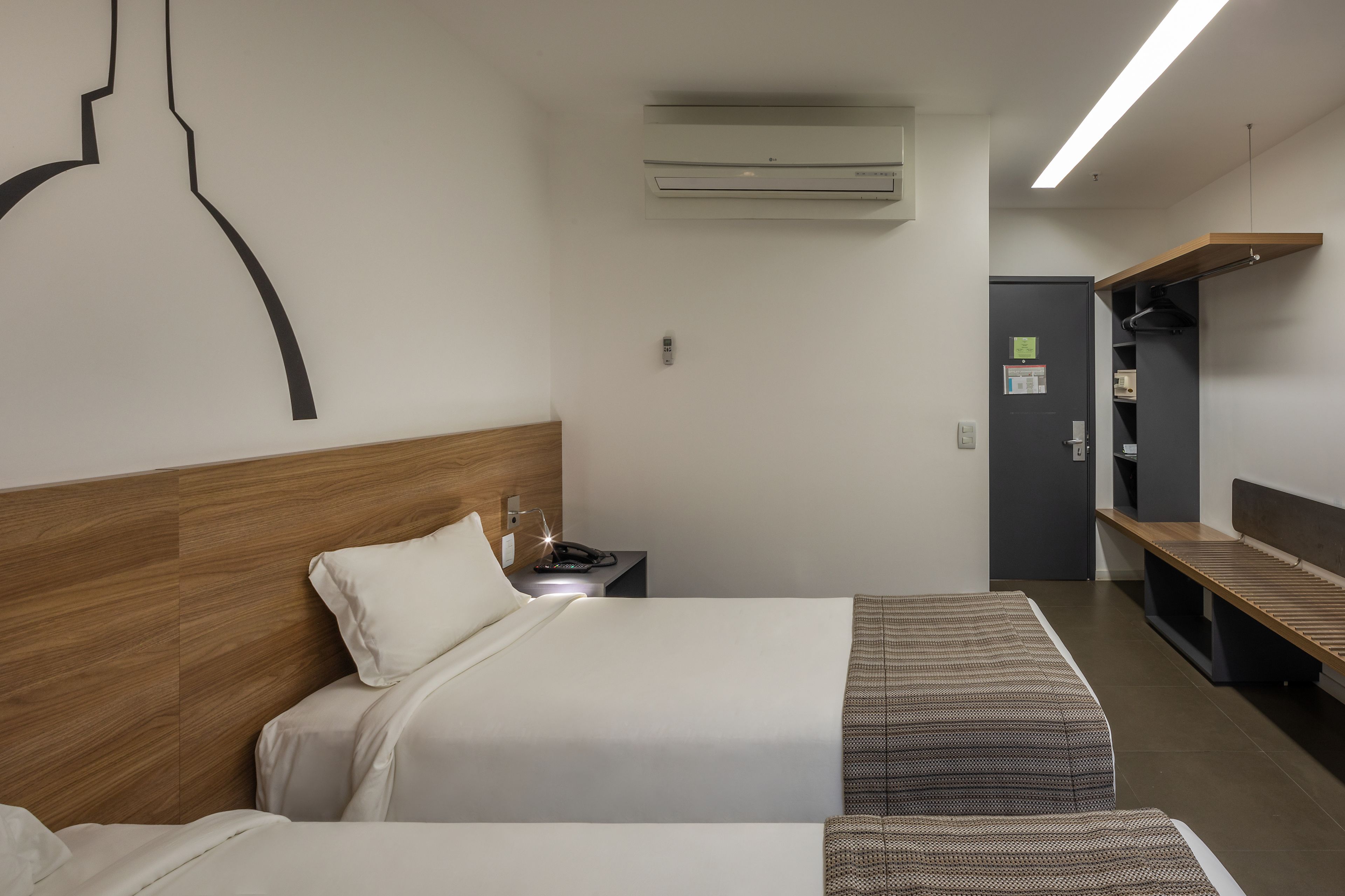 Une chambre d’hôtel avec deux lits, une tête de lit en bois, un bureau avec un téléphone, un appareil de climatisation, un banc et une armoire.