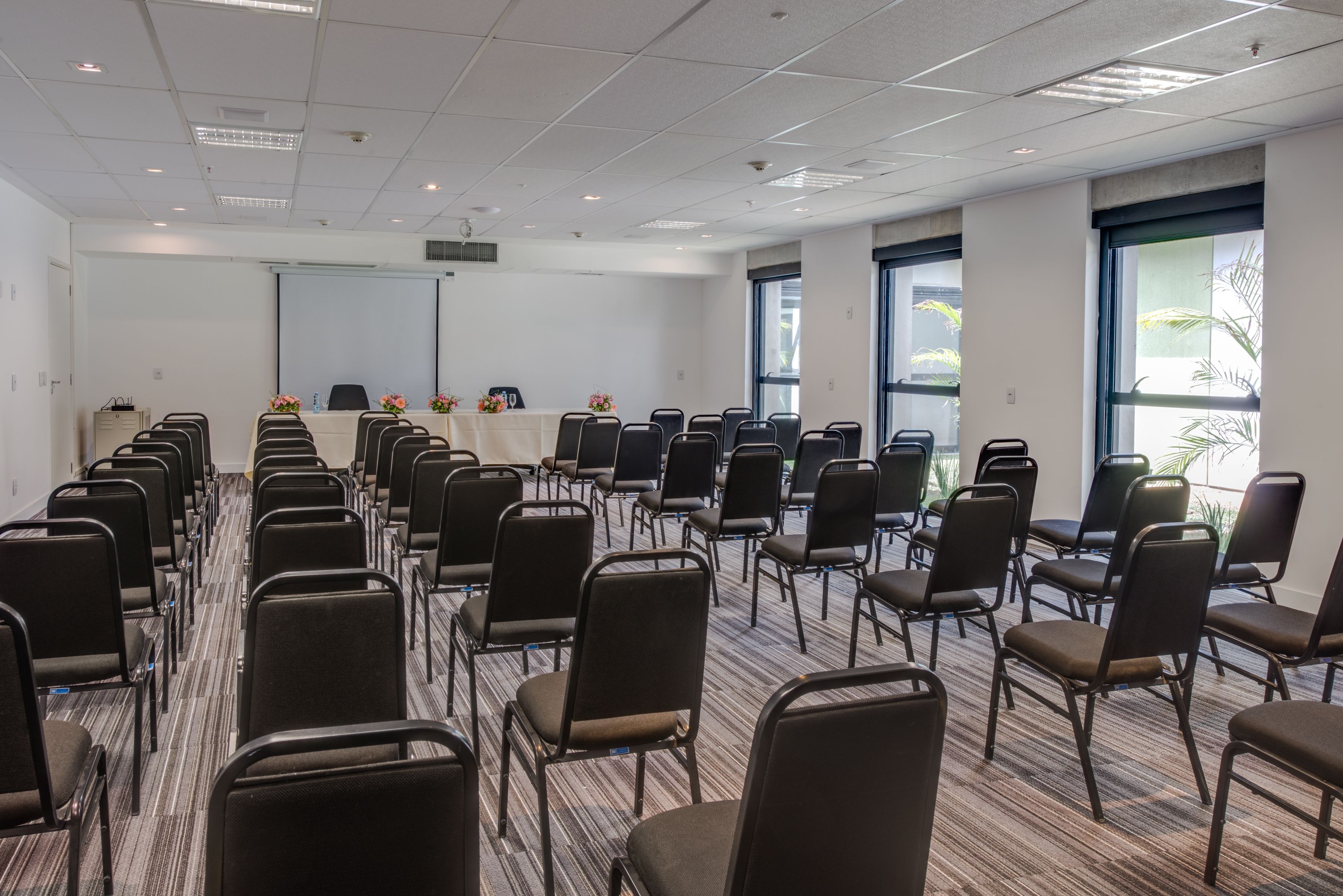 Sala de conferência com fileiras de cadeiras, uma tela de projetor e uma mesa com arranjos florais.